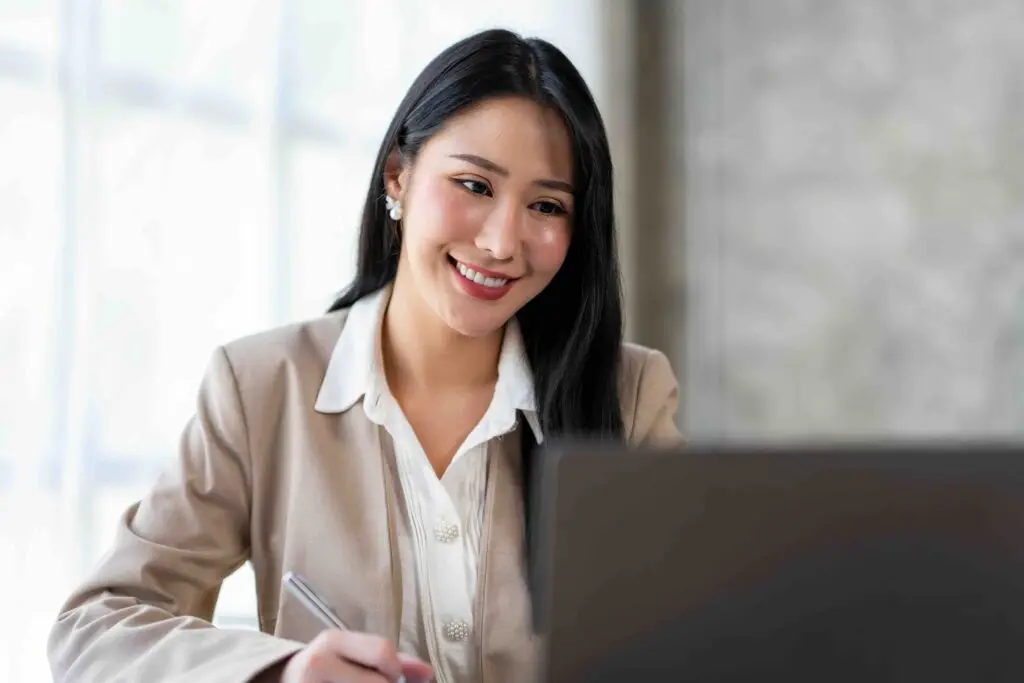 Confident Asian businesswoman sitting and taking notes in modern management concept with laptop computer in office happily.