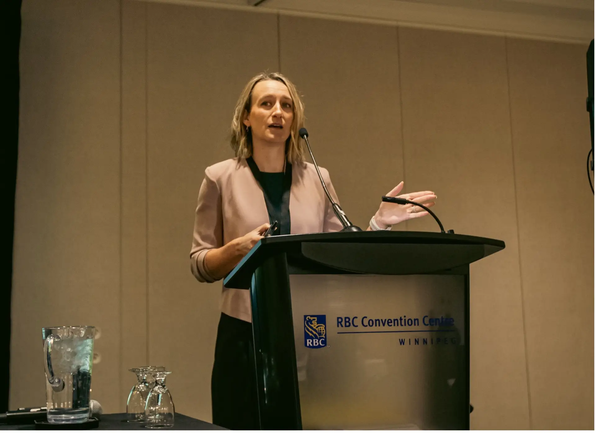 A photo of Dr. Joss Reimer standing before a microphone. A glass jug of water and cups are on a table beside her.